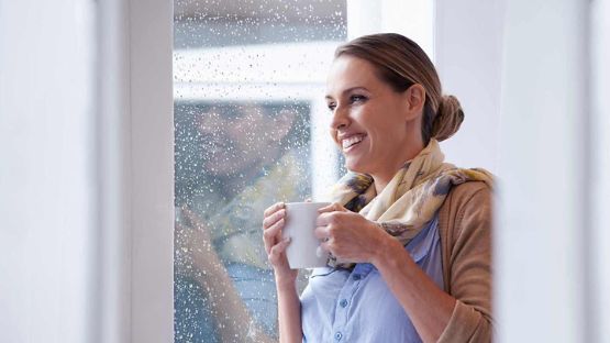 Femme affichant un large sourire et tenant une tasse tout en regardant par la fenêtre par une journée pluvieuse.